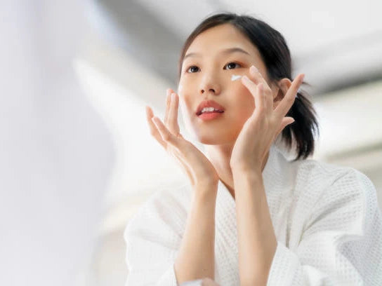 A woman in a bathrobe pressing cream into her cheek in front of a bright window