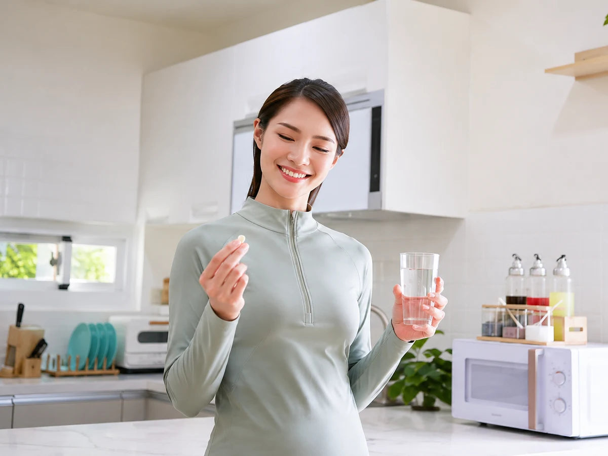 A woman in a bright kitchen with a supplement in one hand and a glass of water in the other