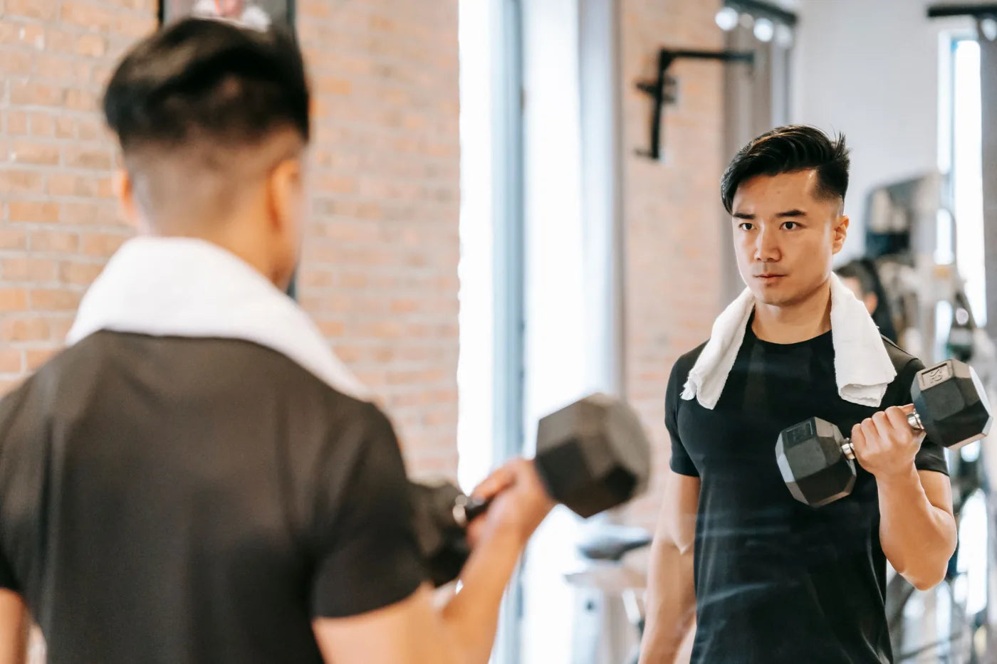 A man training with dumbbells in front of a gym mirror, a towel around his neck